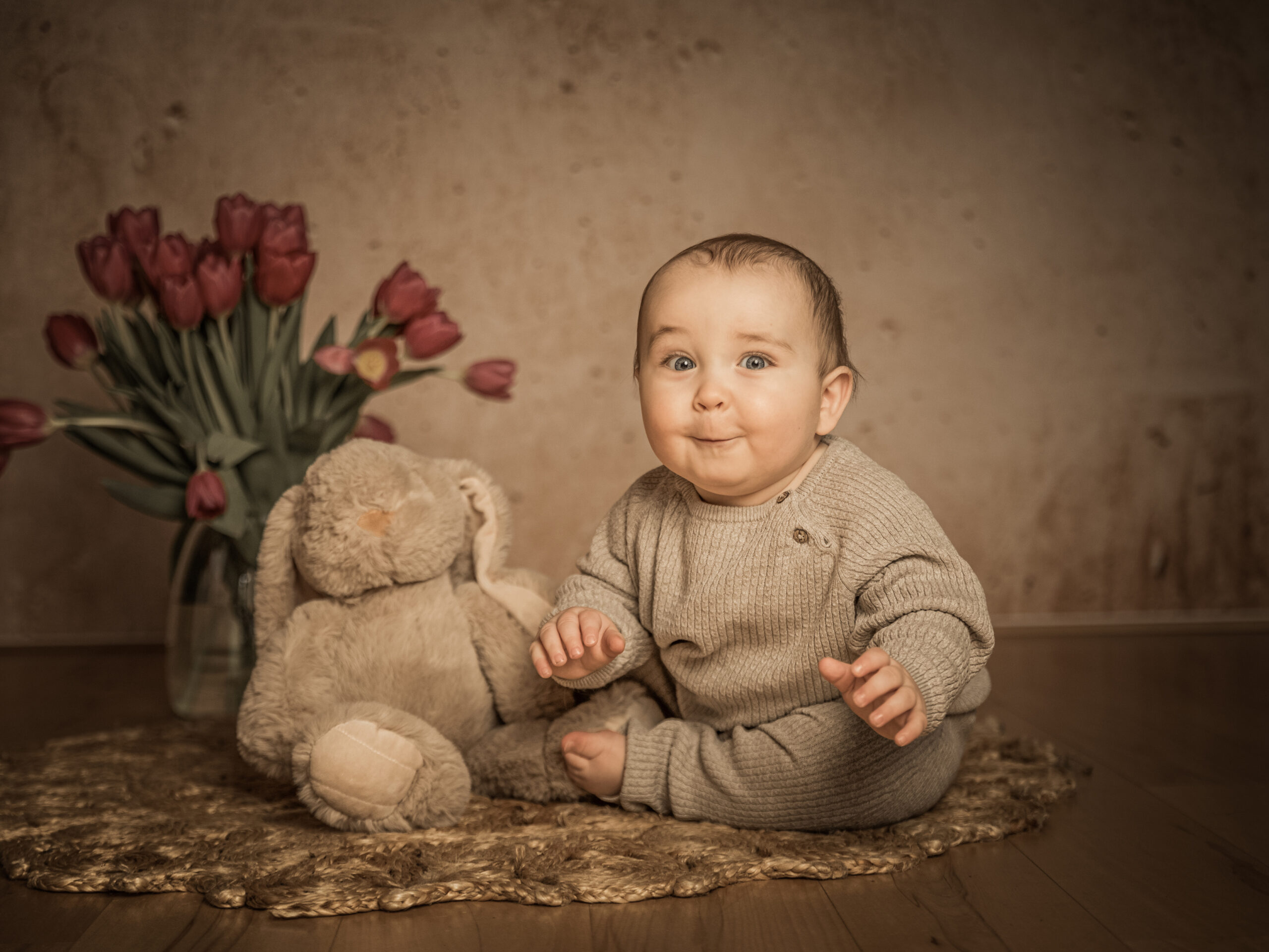 9 Monate altes Baby sitzt auf einem Boho teppich und grinst in die Kamera im Hintergrund steht ein Strauss Tulpen und ein kleiner Teddybär sitzt daneben
