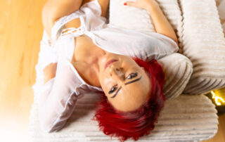 Dessous-Fotografie einer Frau mit weißen Dessous und zarter transparenter Bluse im hellen Backlight auf einem hellen Sofa liegend vor weißem Hintergrund.