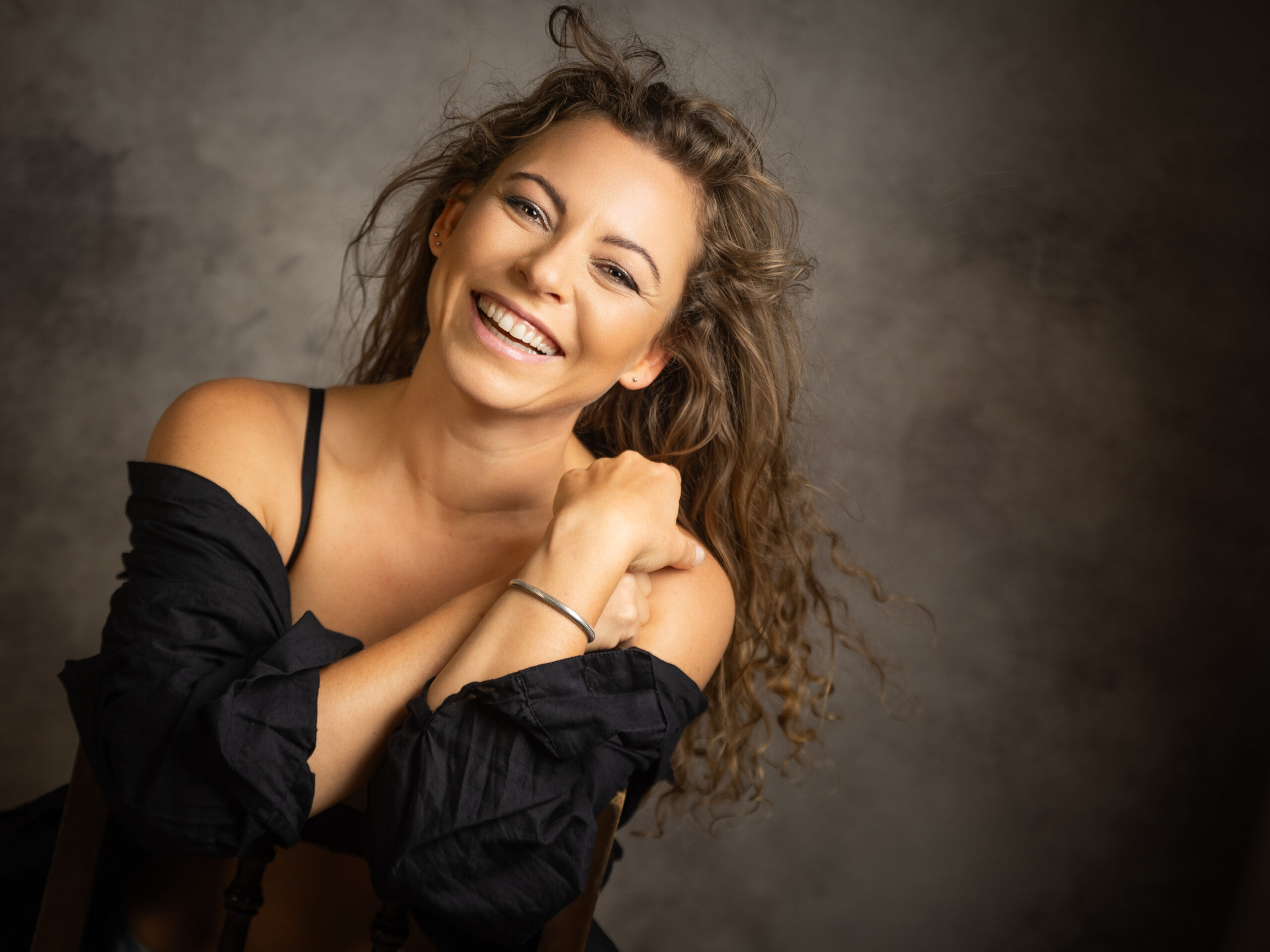 Portraitaufnahme einer Frau, die lächelnd in die Kamera blickt und eine schwarze Bluse trägt, die locker über die Schultern fällt.