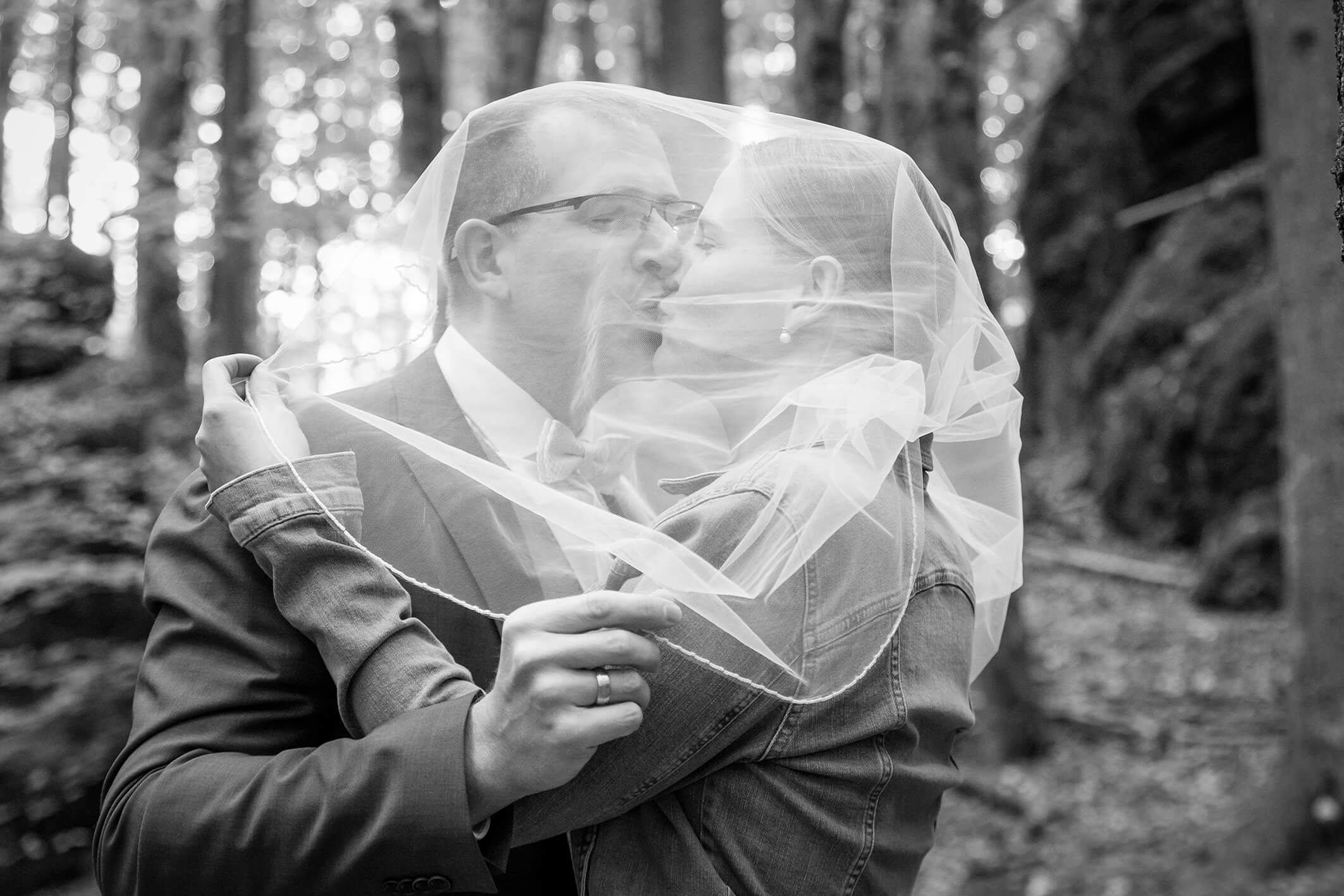Schwarz weiß Portrait eines Brautpaares im Wald, Blätter rahmen das Paar ein, sie küssen sich.