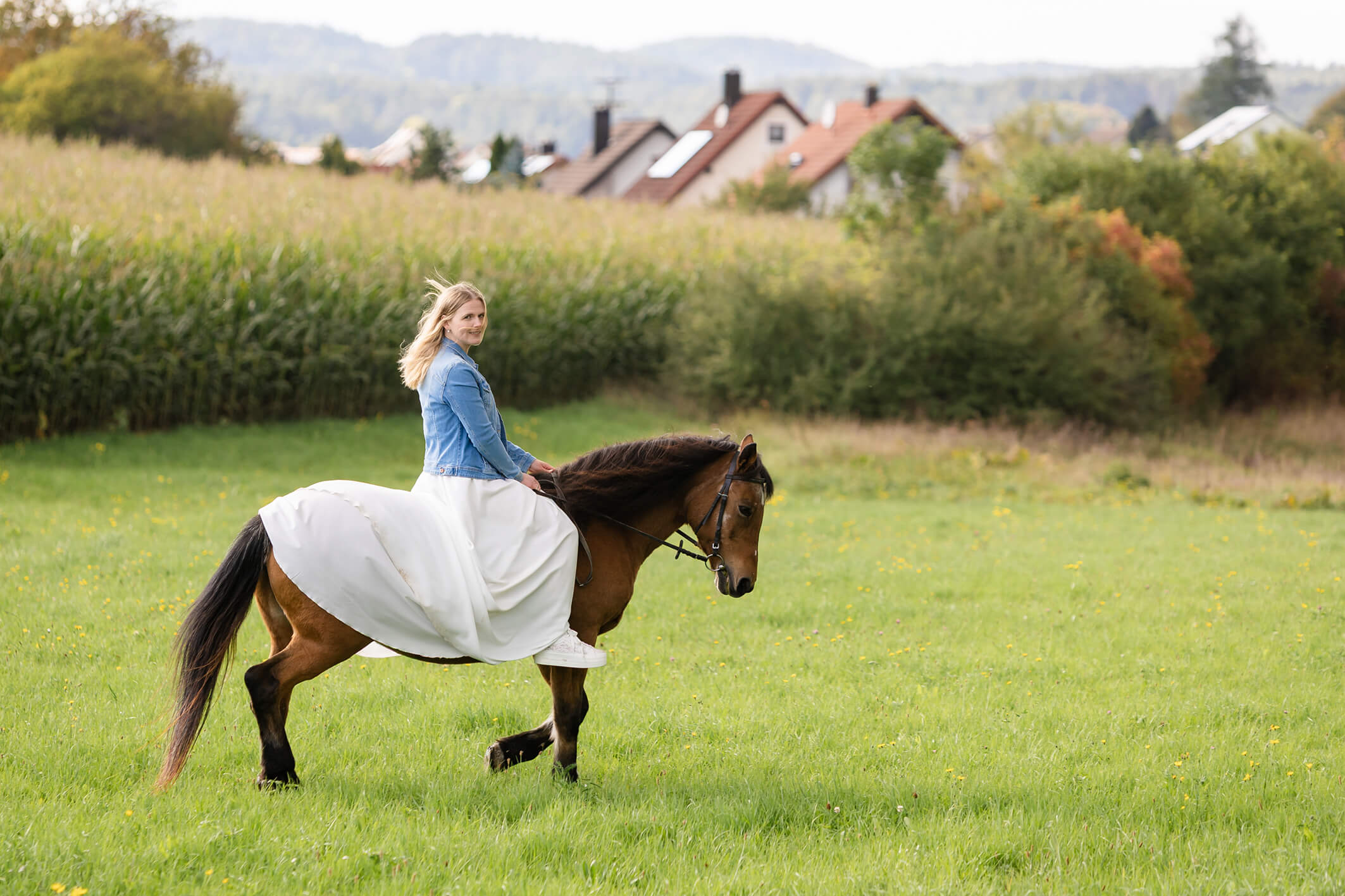 Braut reitet mit Jeansjacke und weißen Turnschuhen auf einem Pferd über die Wiese, moderne Hochzeitsszene.
