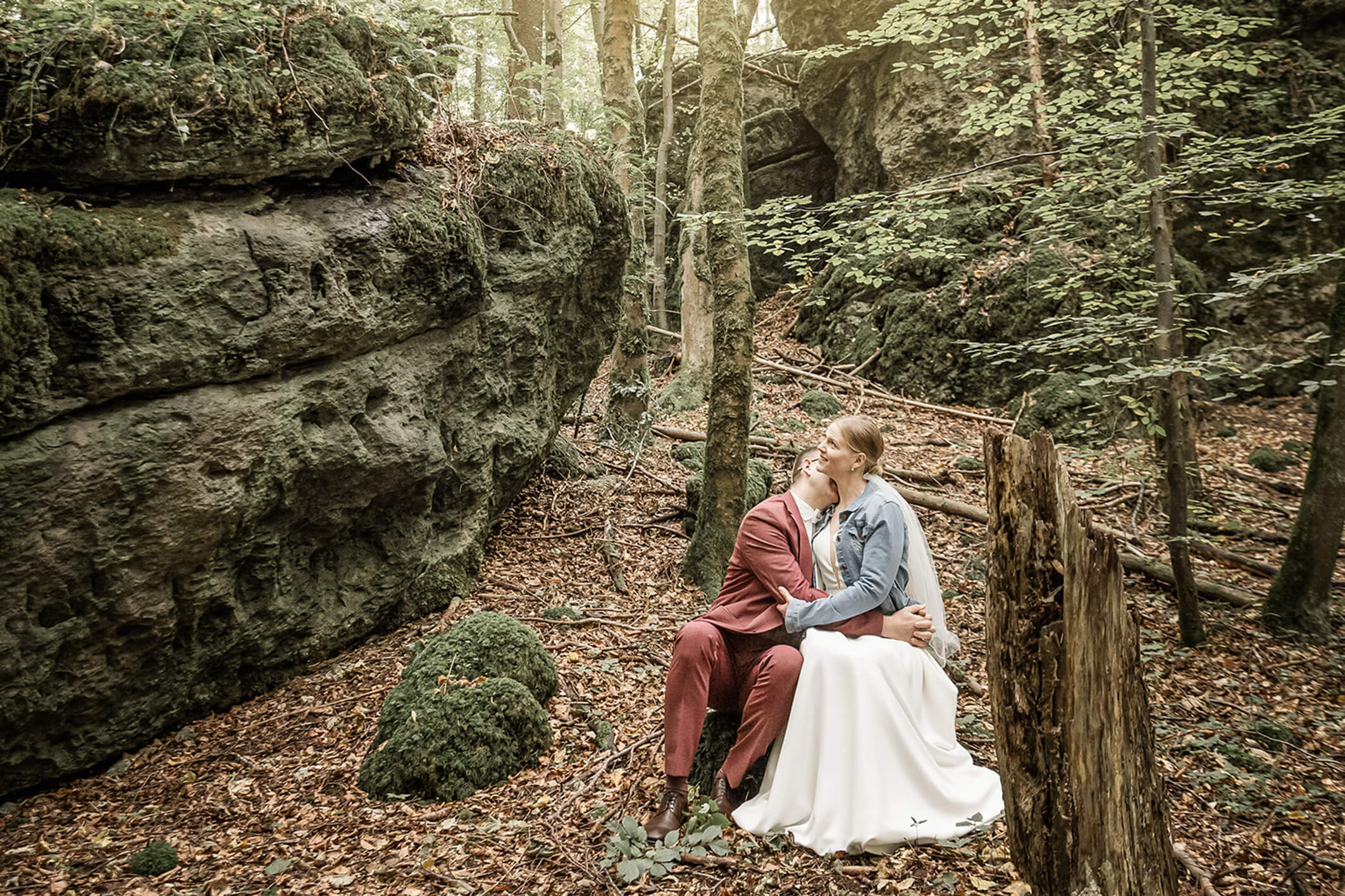 Brautpaar sitzt auf einem Baumstamm im Wald und kuschelt, Sonnenlicht fällt durch die Bäume im Vintage Stil.