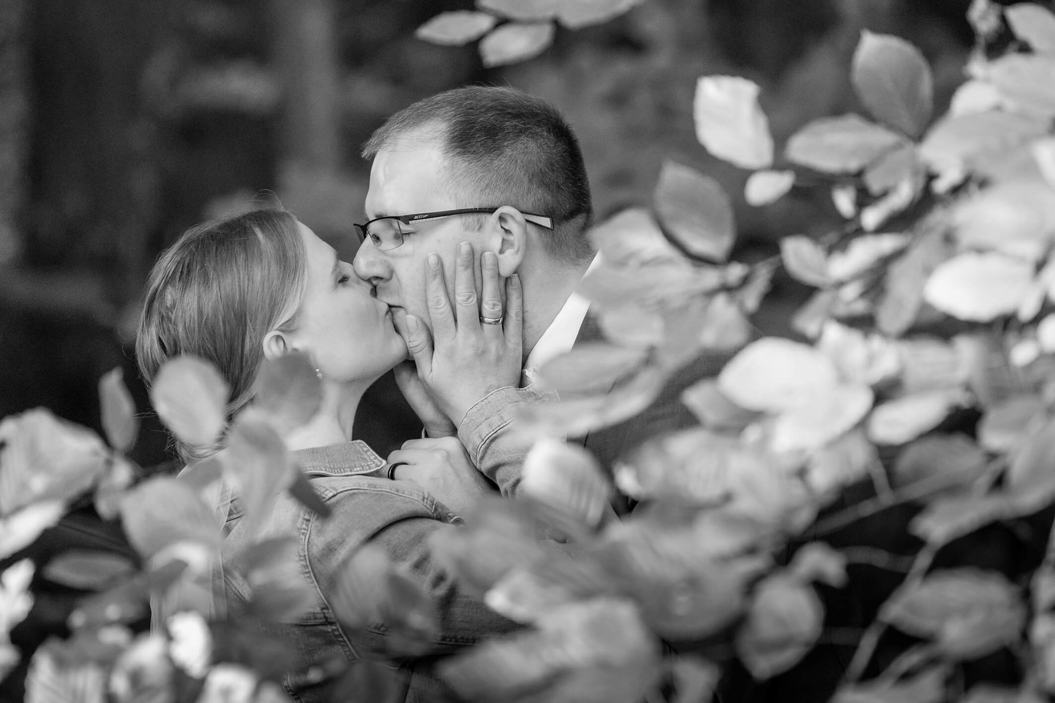 Schwarz weiß Portrait eines Brautpaares im Wald, Blätter rahmen das Paar ein, sie küssen sich.