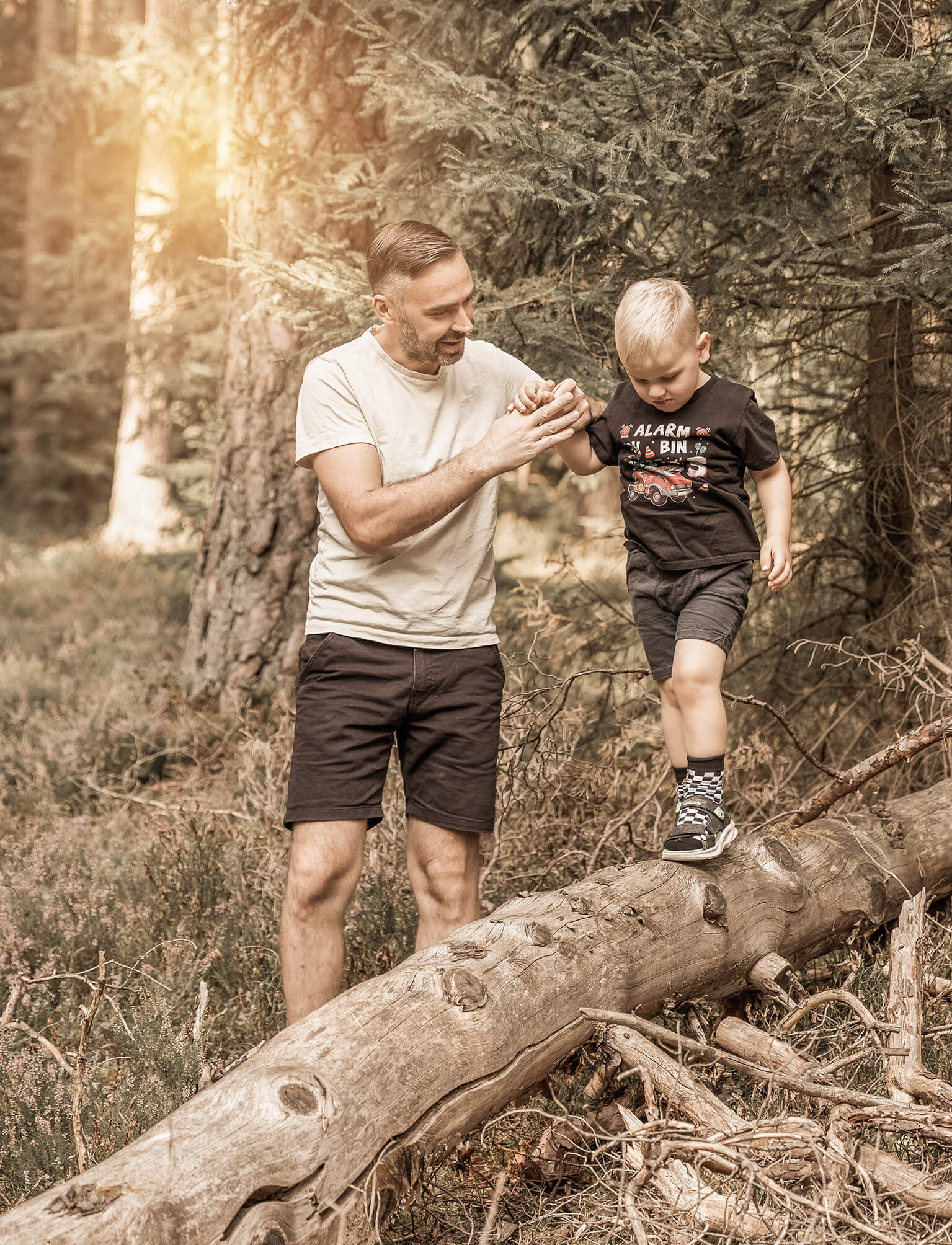 Familienfoto-indernatur-vater-Sohn-doris-dörfler-pegnitz Vater und Sohn im Wald, der Junge balanciert auf einem Baumstamm im Sonnenlicht.