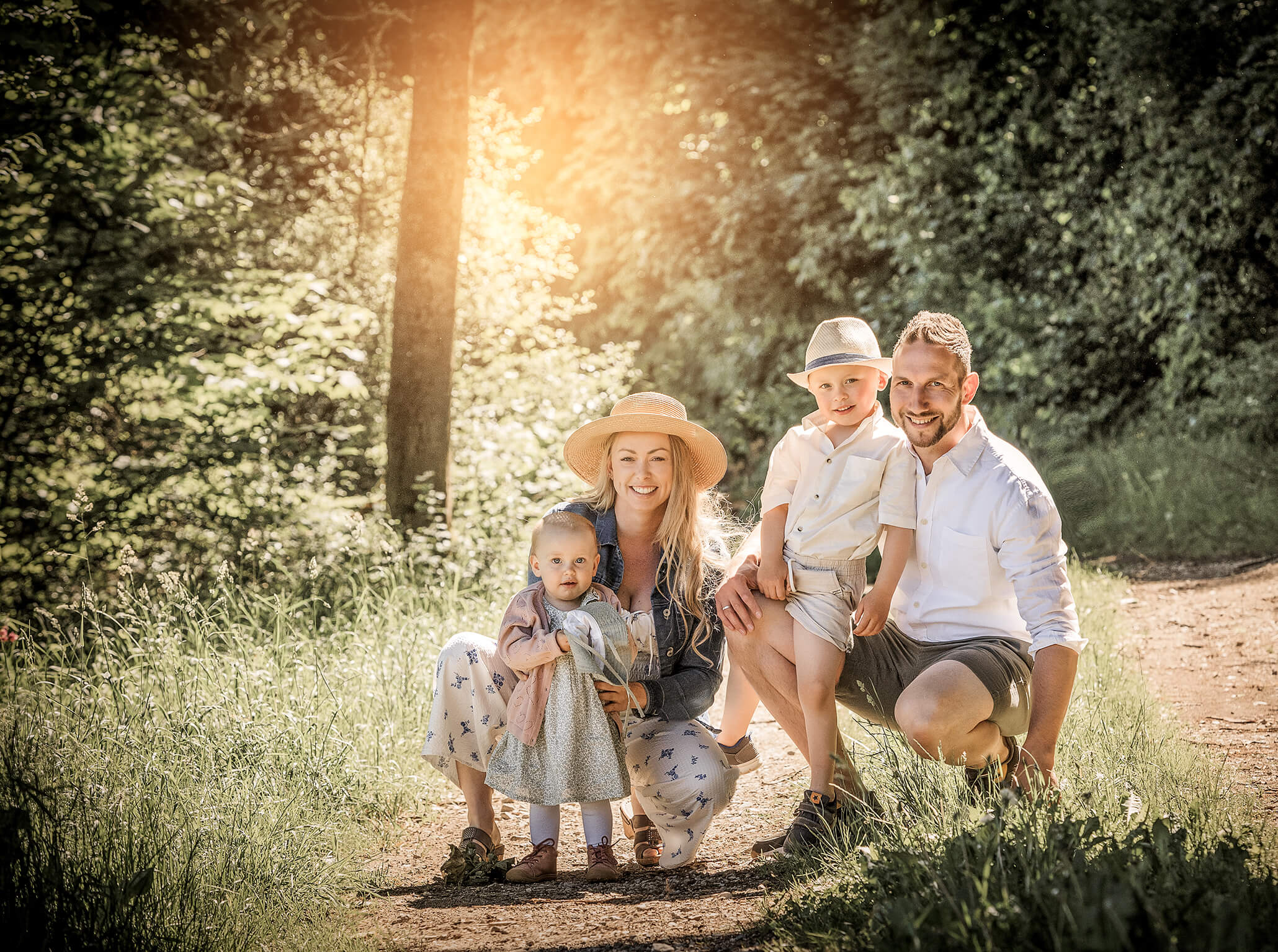 amschlossber-pegnitz-fränkischeschweiz-doris-dörfler-farbenfroh-familieylove Familie in der Hocke auf einem Weg in der Natur, mit Hüten und Sonnenlicht im Hintergrund.
